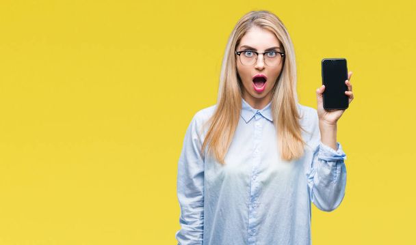 Young beautiful blonde business woman showing screen of smartphone over isolated background scared in shock with a surprise face, afraid and excited with fear expression - Photo, Image