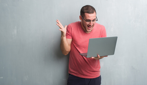 Jeune homme caucasien sur le mur gris grunge en utilisant ordinateur portable très heureux et excité, expression gagnante célébrant la victoire en criant avec un grand sourire et les mains levées
 - Photo, image