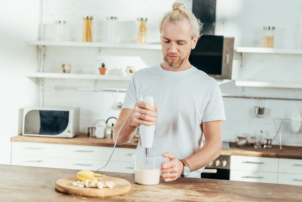 adult man mixing banana protein shake with electric blender at home - Foto, afbeelding