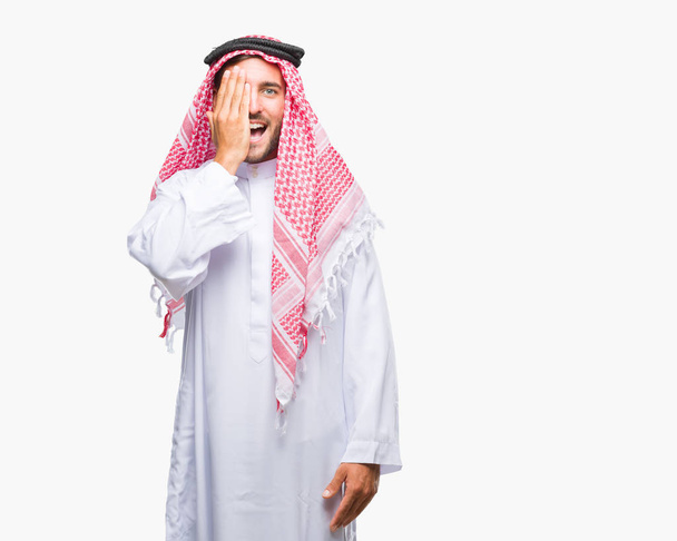 Όμορφος νεαρός που φοράει keffiyeh πέρα από το απομονωμένο υπόβαθρο που καλύπτει ένα μάτι με το χέρι με αυτοπεποίθηση χαμόγελο στο πρόσωπο και έκπληξη συγκίνηση. - Φωτογραφία, εικόνα