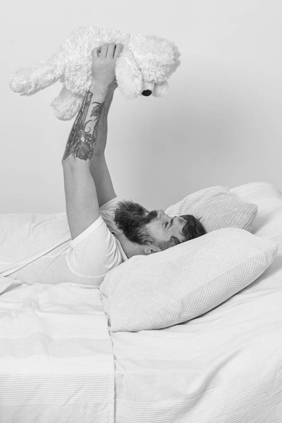 Macho with beard and mustache relaxing with plush toy after nap. Childhood memories concept. Guy on smiling face playing with teddy bear. Man laying on bed and holds toy, white wall on background - Foto, Imagem