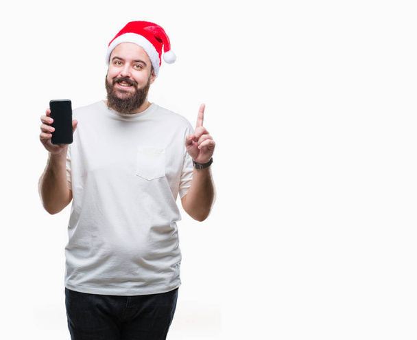 Young caucasian man wearing christmas hat showing smartphone screen over isolated background surprised with an idea or question pointing finger with happy face, number one - Фото, изображение