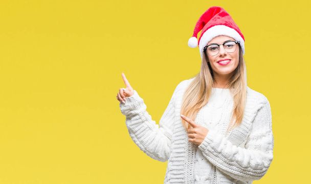 Fiatal gyönyörű nő, fárasztó karácsonyi kalap alatt elszigetelt háttér és mosolyogva néz a kamerába, mutatva a két kéz és az ujjak az oldalon. - Fotó, kép