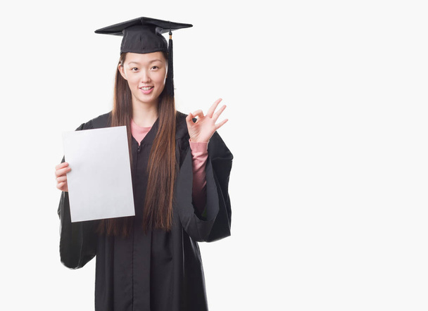 Joven mujer china con uniforme de graduado sosteniendo el grado de papel haciendo signo ok con los dedos, excelente símbolo
 - Foto, Imagen