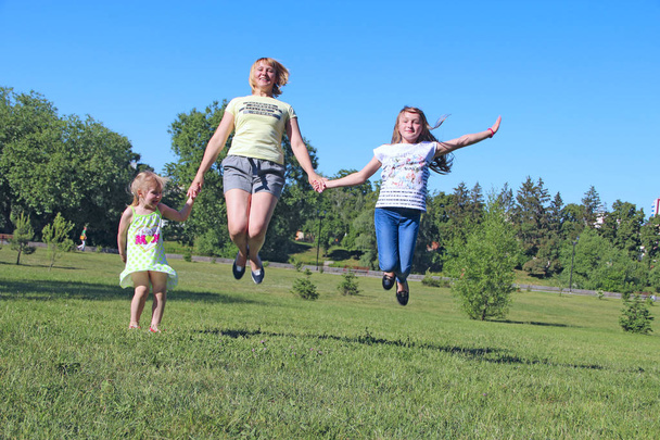 Χαρούμενη μητέρα που διασκεδάζει πηδώντας με τις κόρες της στο πράσινο γρασίδι. Χαρούμενη οικογενειακή ομάδα. Οικογένεια απολαμβάνει τις καλοκαιρινές διακοπές. Άτομα που απολαμβάνουν οικογενειακές διακοπές - Φωτογραφία, εικόνα