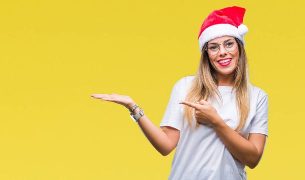 junge schöne Frau mit Weihnachtsmütze über isoliertem Hintergrund erstaunt und lächelnd in die Kamera, während sie mit der Hand präsentiert und mit dem Finger zeigt. - Foto, Bild