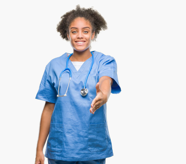 Joven mujer médica afroamericana sobre fondo aislado sonriendo amistoso ofreciendo apretón de manos como saludo y bienvenida. Negocios exitosos
. - Foto, Imagen