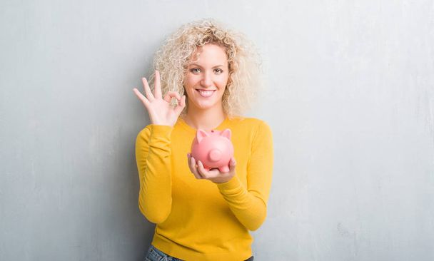 junge blonde Frau über grunge grauen Hintergrund hält Sparschwein tun ok Zeichen mit den Fingern, ausgezeichnetes Symbol - Foto, Bild