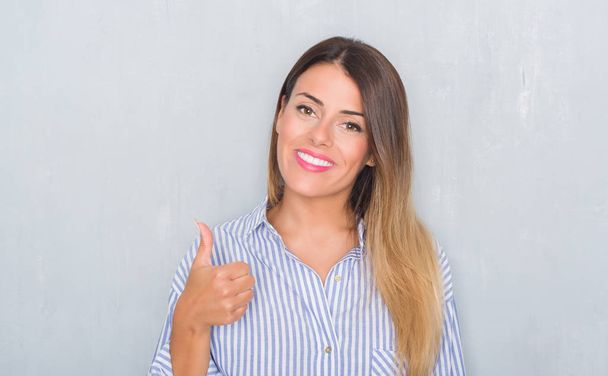 Jonge volwassen vrouw over grunge grijs muur dragen business shirt gelukkig duimschroef opwaarts gebaar met de hand te doen. Goedkeuring van expressie kijken naar de camera met tonen van succes. - Foto, afbeelding
