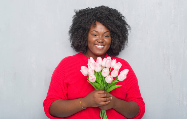 Joven mujer afroamericana sobre pared grunge gris sosteniendo ramo de flores rosadas con una cara feliz de pie y sonriendo con una sonrisa confiada mostrando los dientes
 - Foto, imagen