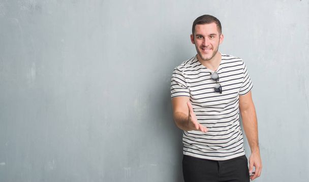 Молодой кавказский человек над серой гранж-стеной улыбается дружелюбно предлагая рукопожатие в качестве приветствия и приветствия. Успешный бизнес
. - Фото, изображение