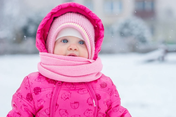 Baby in winter overalls verpakt in een sjaal loopt op een besneeuwde straat en beeldhouwt van sneeuw. - Foto, afbeelding