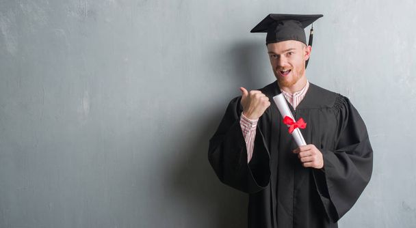 Young redhead man over grey grunge wall wearing graduate uniform holding degree pointing and showing with thumb up to the side with happy face smiling - Φωτογραφία, εικόνα