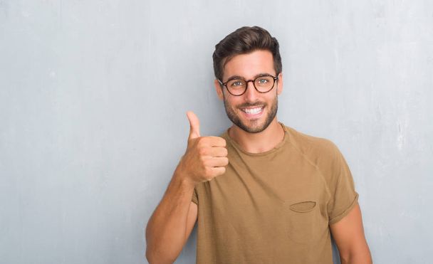 Csinos, fiatal férfi szürke grunge falon szemüveg boldog remek gesztus kézzel csinál. Kifejezés nézte a kamera mutatja a sikeres jóváhagyásáról. - Fotó, kép