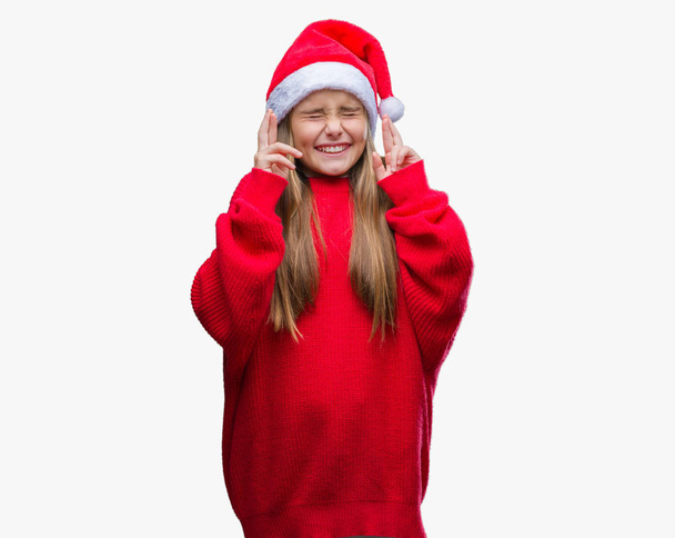 Молодая красивая девушка в рождественской шляпе на изолированном фоне улыбается скрещивая пальцы с надеждой и закрытыми глазами. Удача и суеверие
. - Фото, изображение