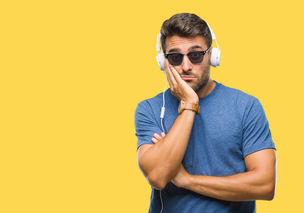 Όμορφος νεαρός φορώντας ακουστικά να ακούτε μουσική μέσω απομονωμένες υπόβαθρο σκέψης ψάχνει κουρασμένη και τρυπημένο με προβλήματα κατάθλιψης με σταυρωμένα χέρια. - Φωτογραφία, εικόνα