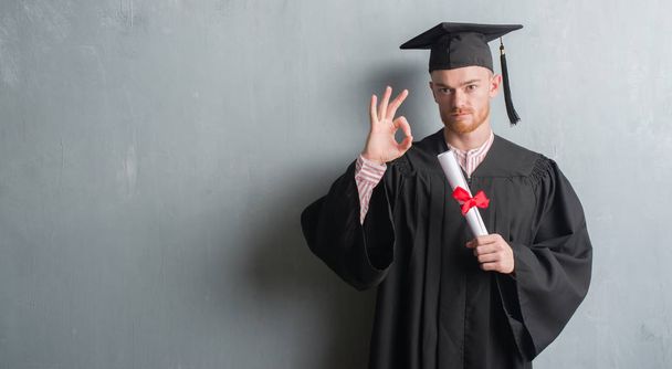 Νέοι κοκκινομάλλα άνδρα πάνω σε γκρι grunge τοίχου φορώντας στολή απόφοιτος κατέχουν πτυχίο κάνει εντάξει πινακίδα με τα δάχτυλα, εξαιρετική σύμβολο - Φωτογραφία, εικόνα