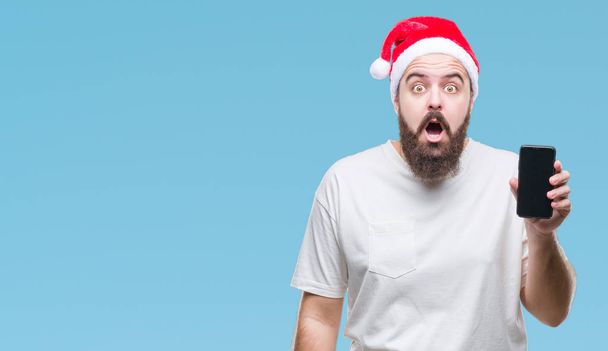 Young caucasian man wearing christmas hat showing smartphone screen over isolated background scared in shock with a surprise face, afraid and excited with fear expression - Foto, Imagem