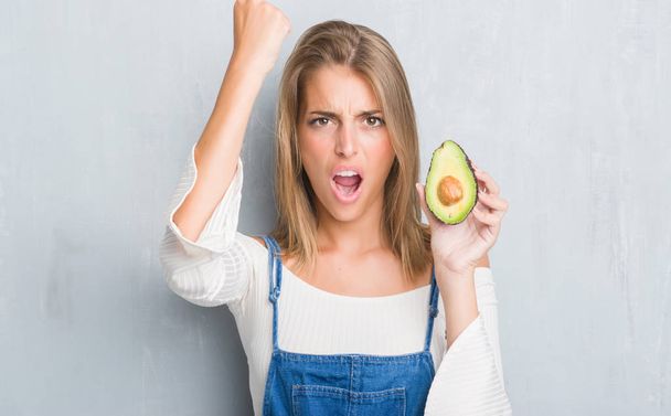 Beautiful young woman over grunge grey wall eating avocado annoyed and frustrated shouting with anger, crazy and yelling with raised hand, anger concept - Photo, Image