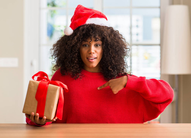 Femme afro-américaine tenant présent portant un chapeau rouge de Noël avec un visage surprise pointant du doigt vers lui-même
 - Photo, image
