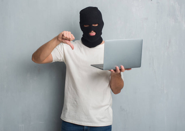Молодой кавказский хакер за серой гранж-стеной делает кибератаку с помощью ноутбука с сердитым лицом, знак негатива показывает неприязнь с опущенными пальцами, концепция отказа
 - Фото, изображение