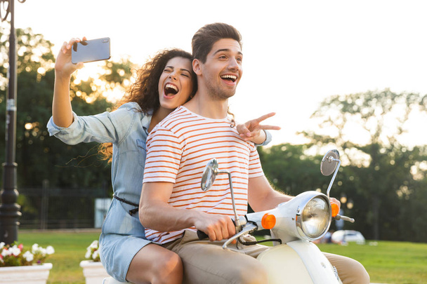 Portrait of cheerful woman taking selfie on smartphone while riding on motorbike through city street together with her boyfriend - Zdjęcie, obraz