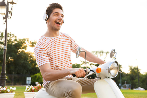 Portrait of joyful man 20s wearing headphones smiling and riding on motorbike through city street - 写真・画像
