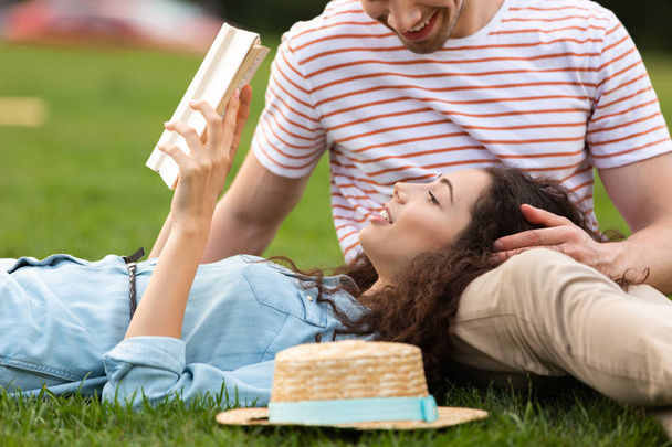 Image of smiling couple man and woman 20s lying on green grass in park and reading book - 写真・画像