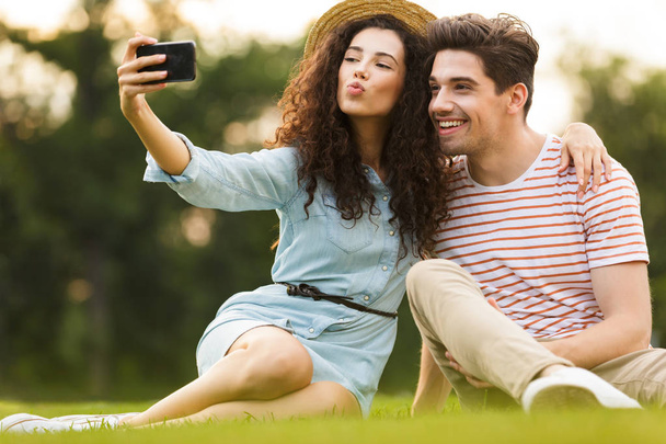 Image of lovely couple man and woman 20s sitting on green grass in park and taking selfie on smartphone - Фото, зображення