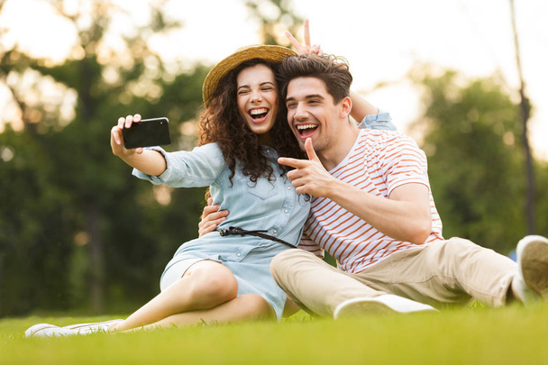 Image of adorable couple man and woman 20s sitting on green grass in park and taking selfie on smartphone - Foto, Imagen