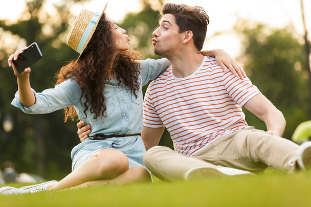 Image of joyful couple man and woman 20s sitting on green grass in park and taking selfie on smartphone - Foto, Imagem