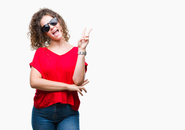 Beautiful brunette curly hair young girl wearing sunglasses over isolated background smiling with happy face winking at the camera doing victory sign. Number two. - Фото, зображення