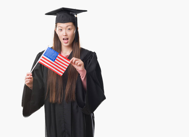 Молодая китаянка в форме выпускницы, держащая в руках флаг Соединенных Штатов Америки, напуганная в шоке с неожиданным лицом, напуганная и взволнованная выражением страха
 - Фото, изображение