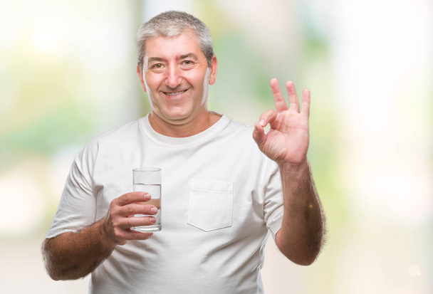 Όμορφος ανώτερος άνθρωπος πίνοντας το ποτήρι νερό πέρα από το απομονωμένο υπόβαθρο κάνει εντάξει πινακίδα με τα δάχτυλα, εξαιρετική σύμβολο - Φωτογραφία, εικόνα