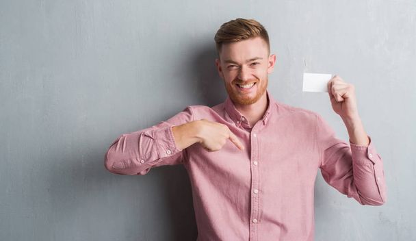 junger rothaariger Mann über graue Grunge-Wand mit leerer Visitenkarte und überraschendem Gesicht, das mit dem Finger auf sich selbst zeigt - Foto, Bild