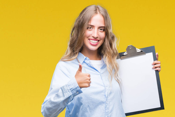 Beautiful young blonde business woman holding clipboard over isolated background happy with big smile doing ok sign, thumb up with fingers, excellent sign - Fotoğraf, Görsel