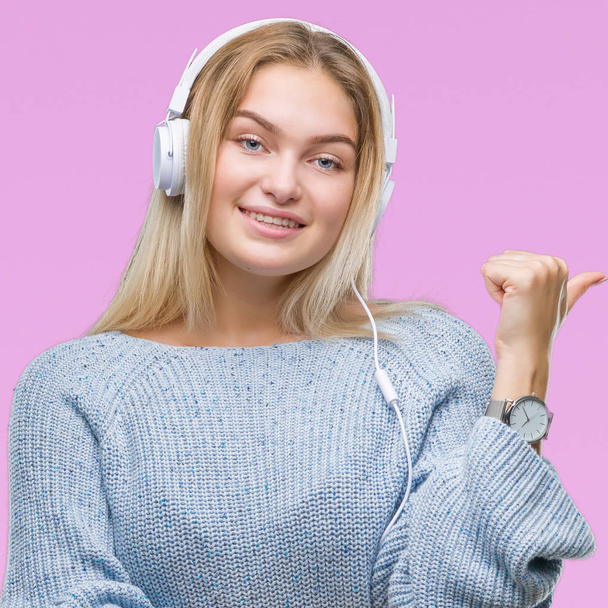 Mujer caucásica joven escuchando música usando auriculares sobre un fondo aislado sonriendo con la cara feliz mirando y apuntando hacia un lado con el pulgar hacia arriba
. - Foto, Imagen