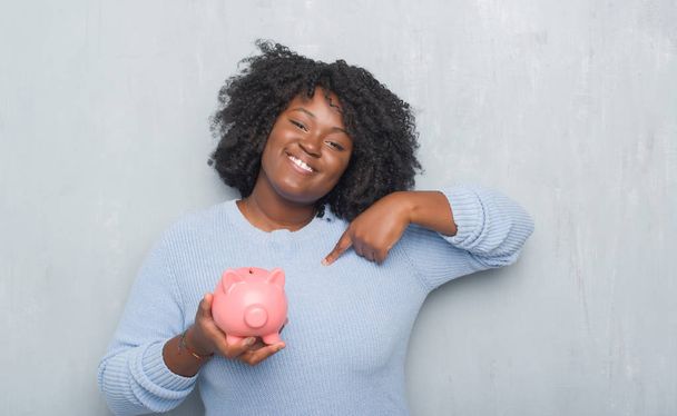 Young african american woman over grey grunge wall holding piggy bank with surprise face pointing finger to himself - Photo, Image