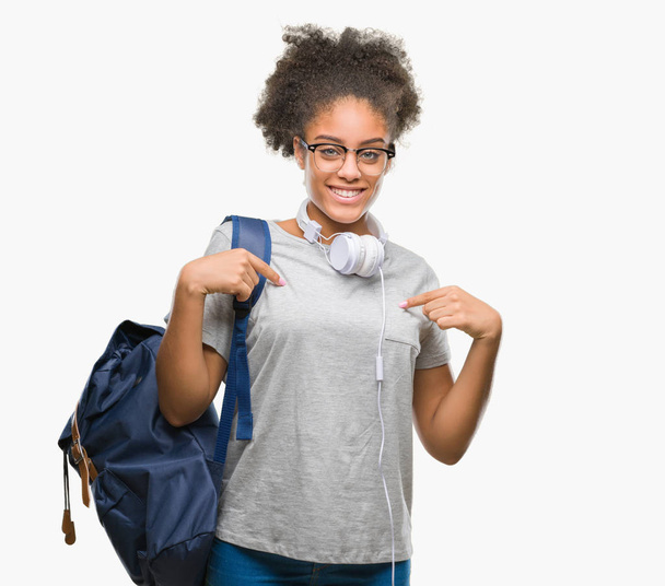 Jovem afro-americana estudante mulher usando fones de ouvido e mochila sobre fundo isolado olhando confiante com sorriso no rosto, apontando-se com os dedos orgulhosos e felizes
. - Foto, Imagem