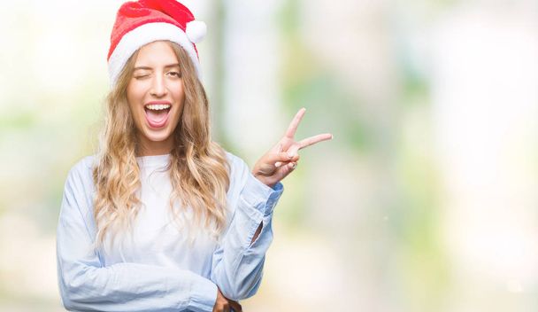 Belle jeune femme blonde portant un chapeau de Noël sur fond isolé souriant avec un clin d'œil heureux à la caméra faisant signe de victoire. Numéro deux
. - Photo, image