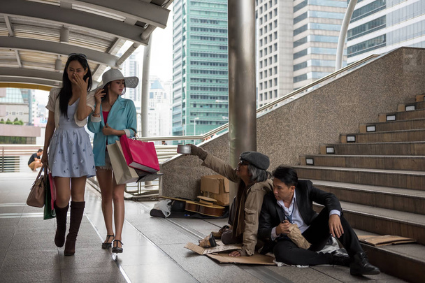 Asian tourist women with many shopping bag look down on smell homeless dirty old guy and drunk businessman in urban city. bad habit for high society or gentility to disgust the poor. - Photo, Image