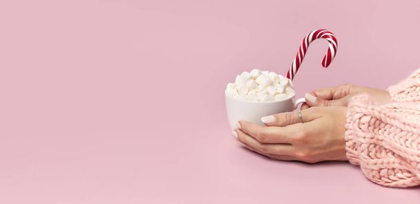 Female hands in knitted sweater holding cup of marshmallows and Christmas candy cane on pink background Flat Lay copy space Winter traditional food Festive decor celebration presents Xmas holiday. - Foto, afbeelding