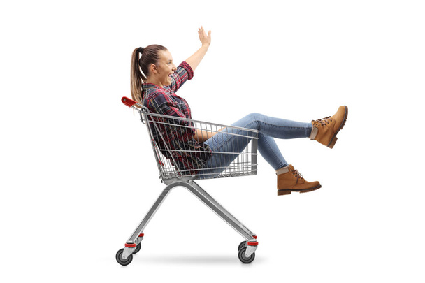Jovem mulher sentada dentro de um carrinho de compras e acenando isolada no fundo branco
 - Foto, Imagem