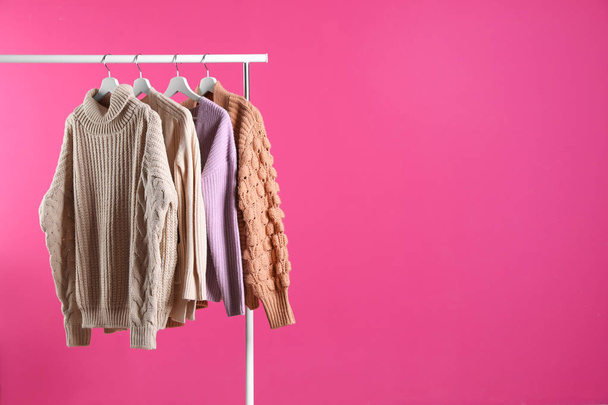 Collection of warm sweaters hanging on rack against color background. Space for text - Фото, изображение