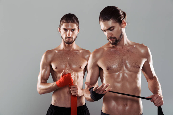 Portrait of a two confident muscular shirtless twin brothers standing isolated over gray background, wrapping hands with boxing wraps - Фото, зображення