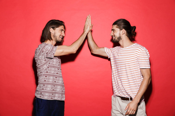 Portrait of two smiling twin brothers standing isolated over red background, giving high five - Fotoğraf, Görsel