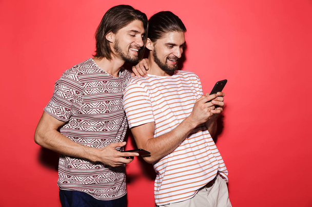携帯電話を使用して、赤の背景に分離された双子の兄弟を笑っている 2 人の若いの肖像画 - 写真・画像