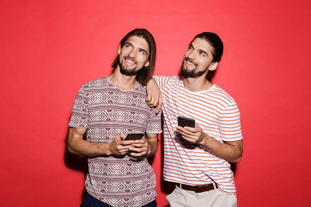 Portrait of a two young smiling twin brothers isolated over red background, using mobile phones, looking up - 写真・画像