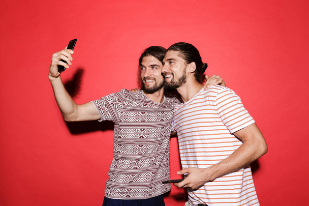 Portrait of a two young smiling twin brothers isolated over red background, using mobile phones, taking a selfie - Foto, imagen