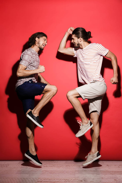 Full length portrait of a two young happy twin brothers isolated over red background, celebrating success, jumping - Zdjęcie, obraz
